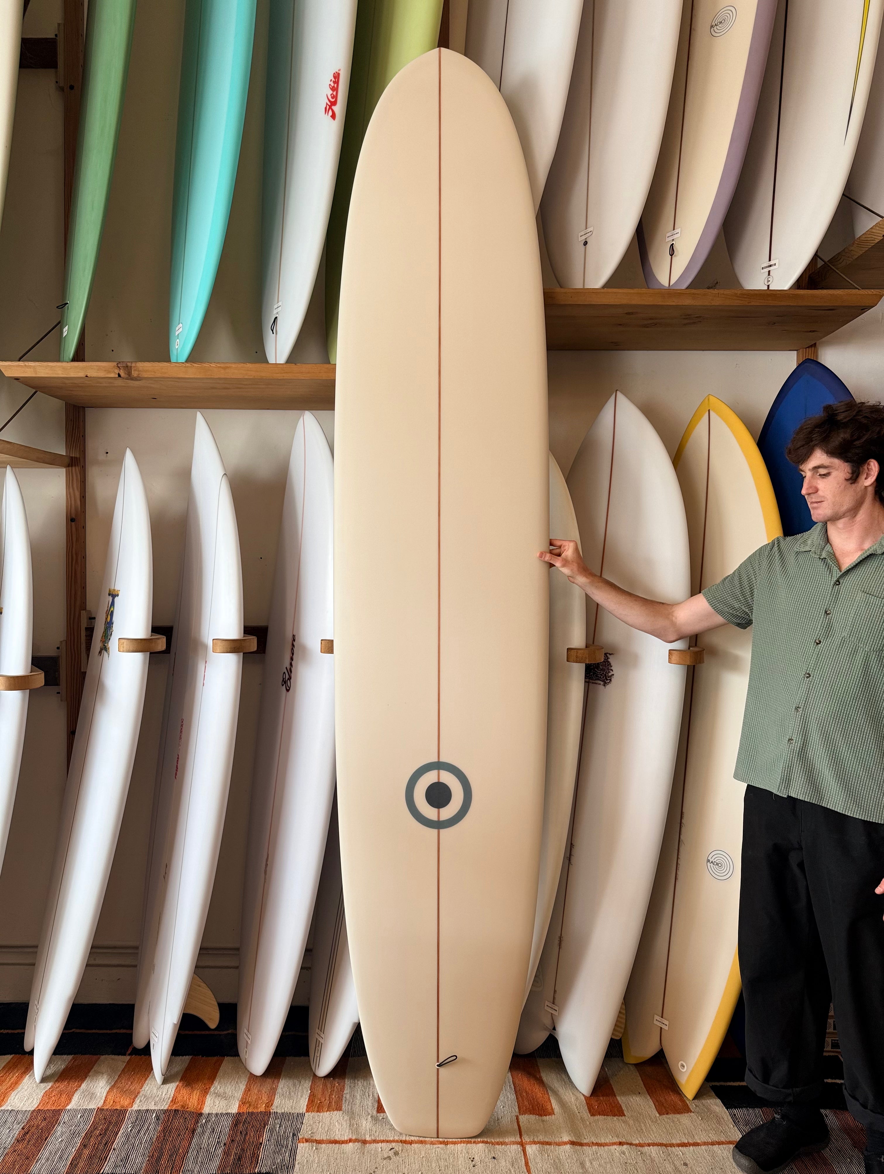 A man in a green shirt and black pants stands next to a cream-colored surfboard with a circular target logo, holding its edge in a shop filled with other surfboards on wooden shelves.