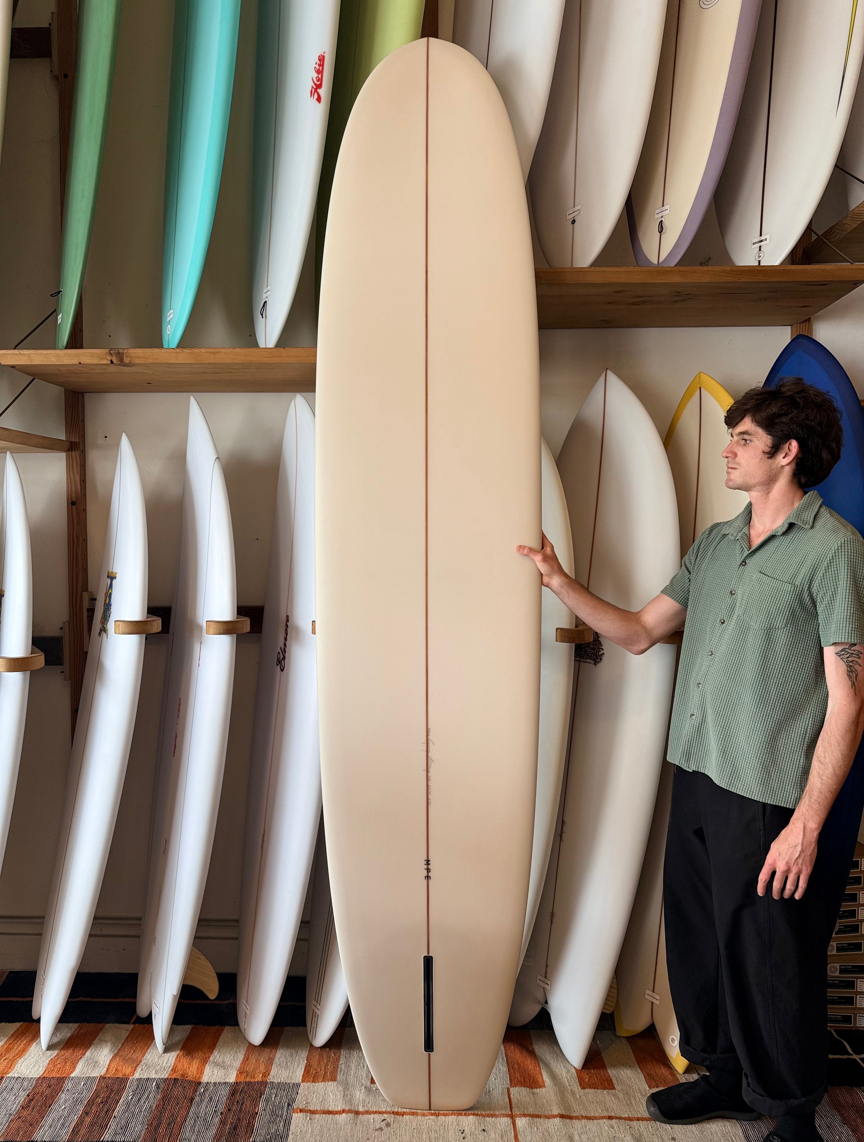 A man in a sage green shirt and black pants stands next to a tall, cream-colored surfboard in a shop filled with other surfboards on wooden shelves.